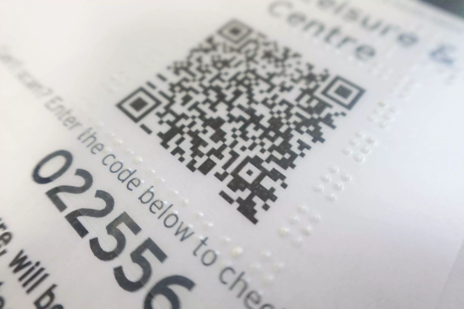 Close-up photograph of a printed sign. A QR code is surrounded by a frame of small raised clear dots that can be felt by touch, with braille alongside. Beneath the code is the printed text “Can’t scan? Enter the code below…” followed by a large reference number.